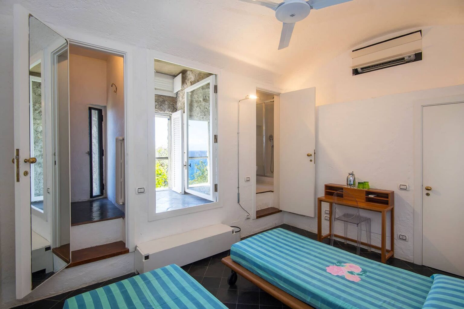 Bright and airy bedroom featuring two twin beds with blue striped linens, a wooden nightstand, and a ceiling fan. The room includes large windows with a scenic view, a mirror, and modern air conditioning, creating a comfortable and inviting atmosphere. | Only on SopranoVillas