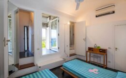 Bright and airy bedroom featuring two twin beds with blue striped linens, a wooden nightstand, and a ceiling fan. The room includes large windows with a scenic view, a mirror, and modern air conditioning, creating a comfortable and inviting atmosphere.