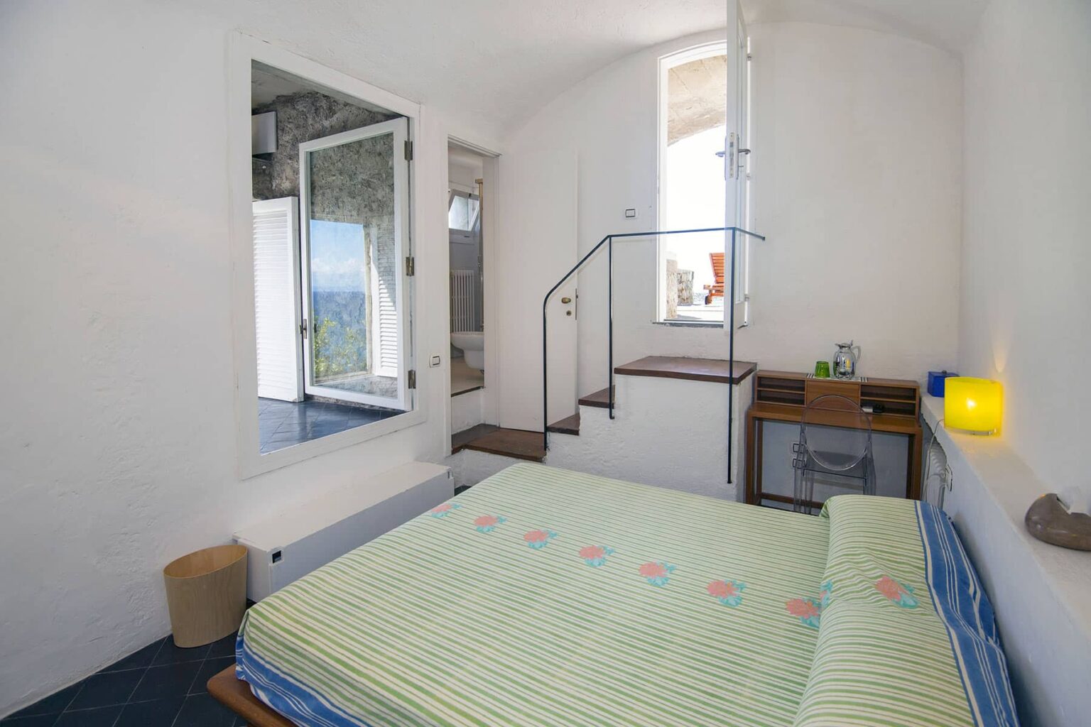 Cozy bedroom featuring a double bed with a green striped blanket, wooden furniture, and a staircase leading to an upper level. The room has large windows allowing natural light, creating a bright and inviting atmosphere. | Only on SopranoVillas