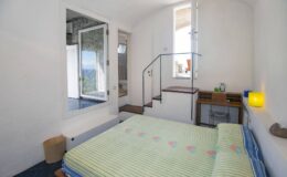 Cozy bedroom featuring a double bed with a green striped blanket, wooden furniture, and a staircase leading to an upper level. The room has large windows allowing natural light, creating a bright and inviting atmosphere.