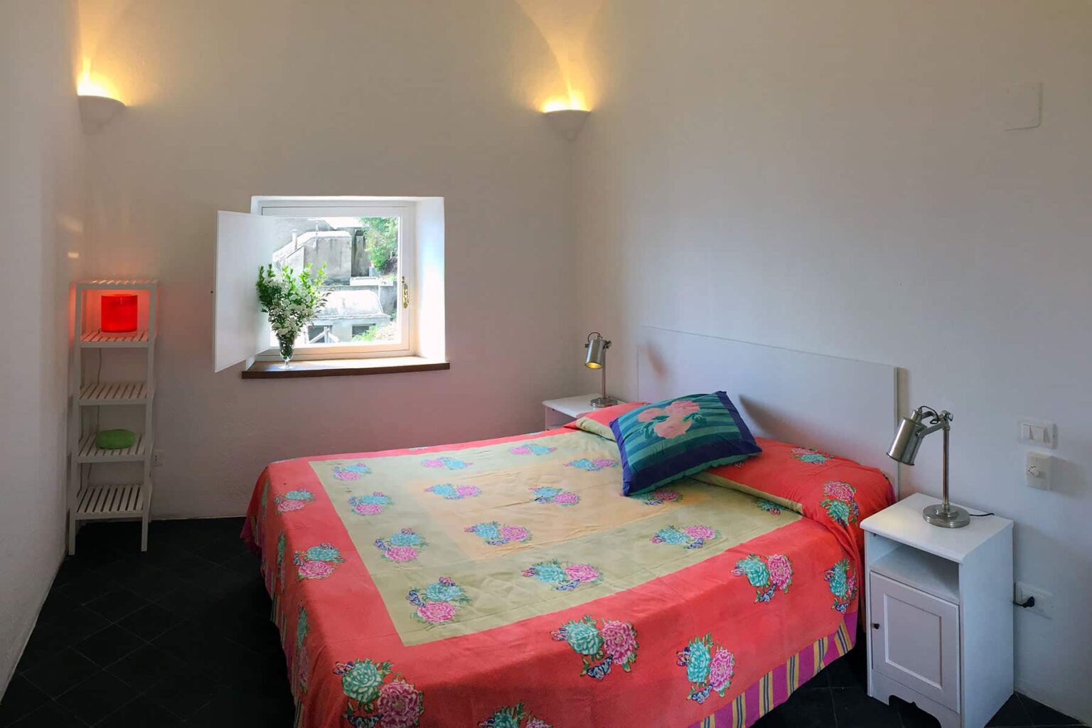 Cozy bedroom featuring a colorful floral bedspread, bedside tables with lamps, and a window with natural light illuminating the space. Ideal for relaxation and comfort. | Only on SopranoVillas