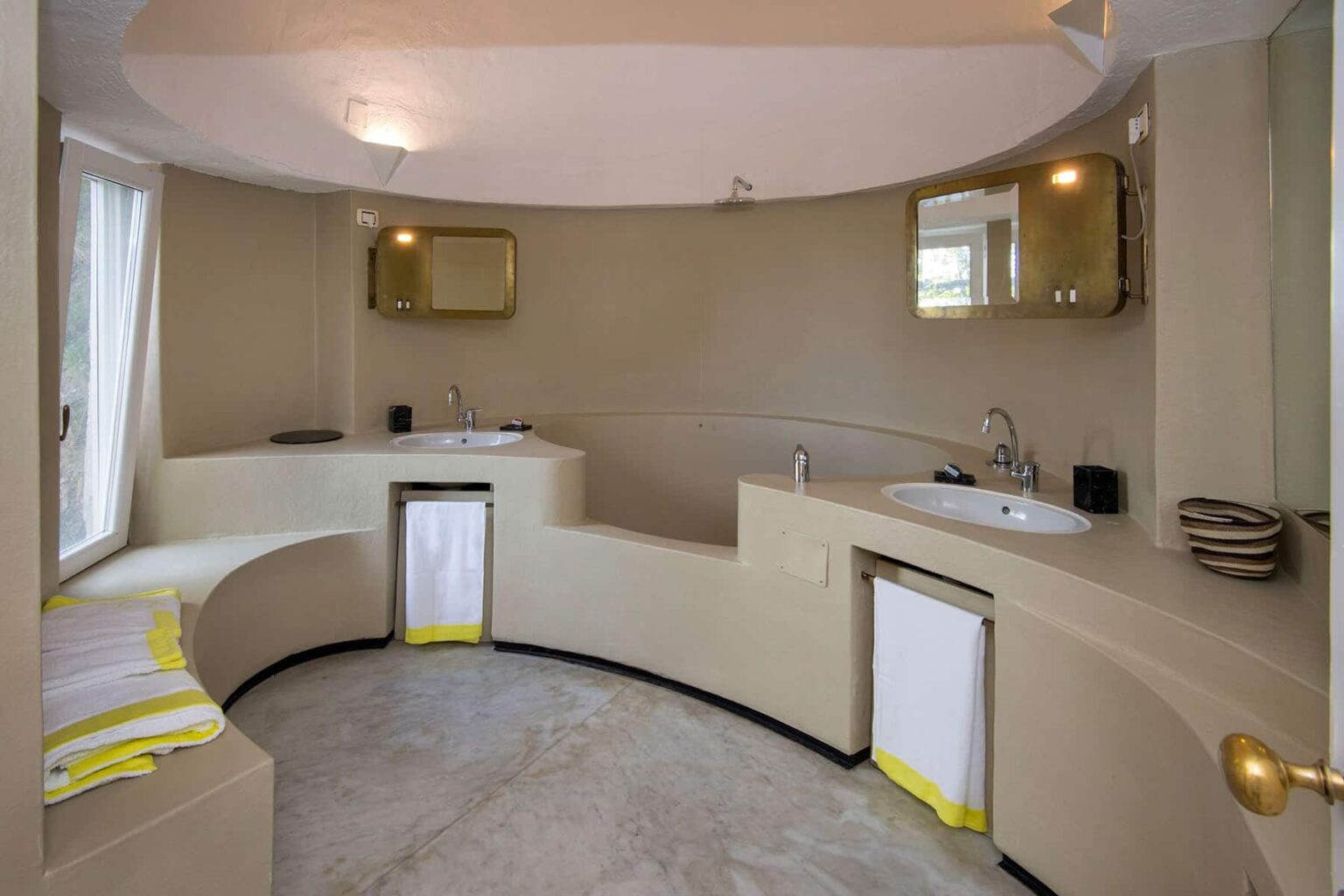 Modern curved bathroom design featuring dual sinks, minimalist decor, and natural light from a window. The walls and fixtures are in neutral tones, complemented by vibrant yellow towels for a touch of color. | Only on SopranoVillas