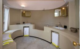Modern curved bathroom design featuring dual sinks, minimalist decor, and natural light from a window. The walls and fixtures are in neutral tones, complemented by vibrant yellow towels for a touch of color.