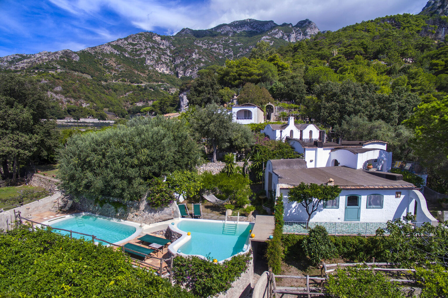 Aerial view of a serene countryside villa featuring two swimming pools surrounded by lush greenery and mountains in the background, showcasing a peaceful retreat atmosphere. | Only on SopranoVillas