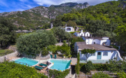 Aerial view of a serene countryside villa featuring two swimming pools surrounded by lush greenery and mountains in the background, showcasing a peaceful retreat atmosphere.