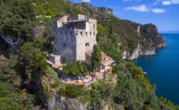 Historic stone tower overlooking the coastline, surrounded by lush greenery and offering a panoramic view of the sea, ideal for vacation rentals or photography.