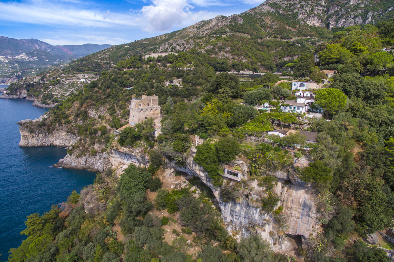Aerial view of a coastal landscape featuring lush greenery and a historic stone tower perched on a cliff overlooking the blue sea, with scenic mountains in the background. | Only on SopranoVillas