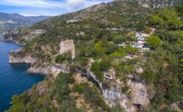 Aerial view of a coastal landscape featuring lush greenery and a historic stone tower perched on a cliff overlooking the blue sea, with scenic mountains in the background.