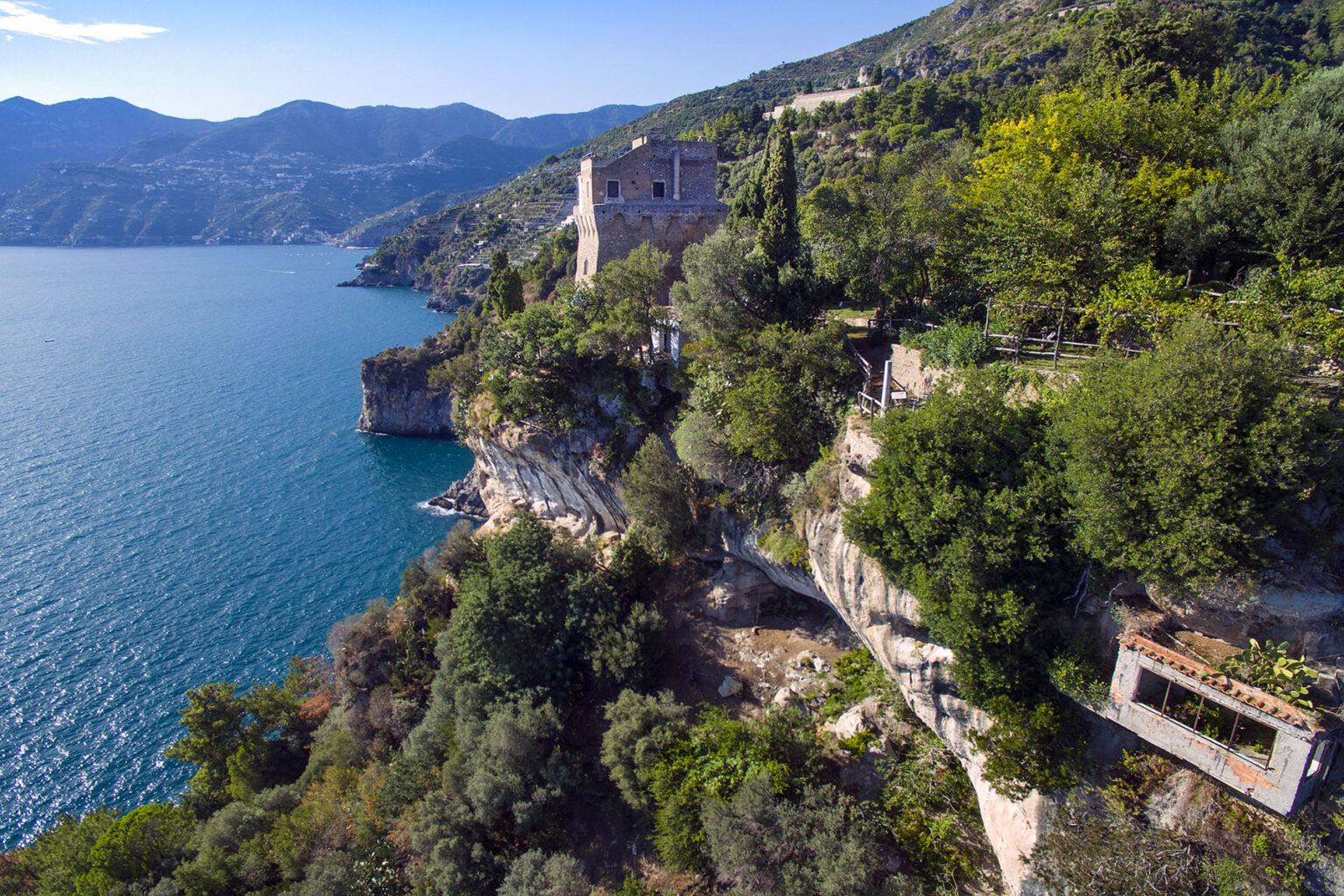 Aerial view of a scenic coastal landscape featuring a charming house perched on a rocky cliff overlooking a serene blue lake, surrounded by lush greenery and mountains in the background. | Only on SopranoVillas