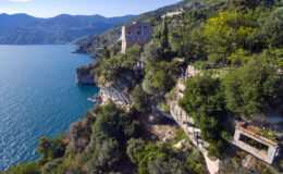 Aerial view of a scenic coastal landscape featuring a charming house perched on a rocky cliff overlooking a serene blue lake, surrounded by lush greenery and mountains in the background.