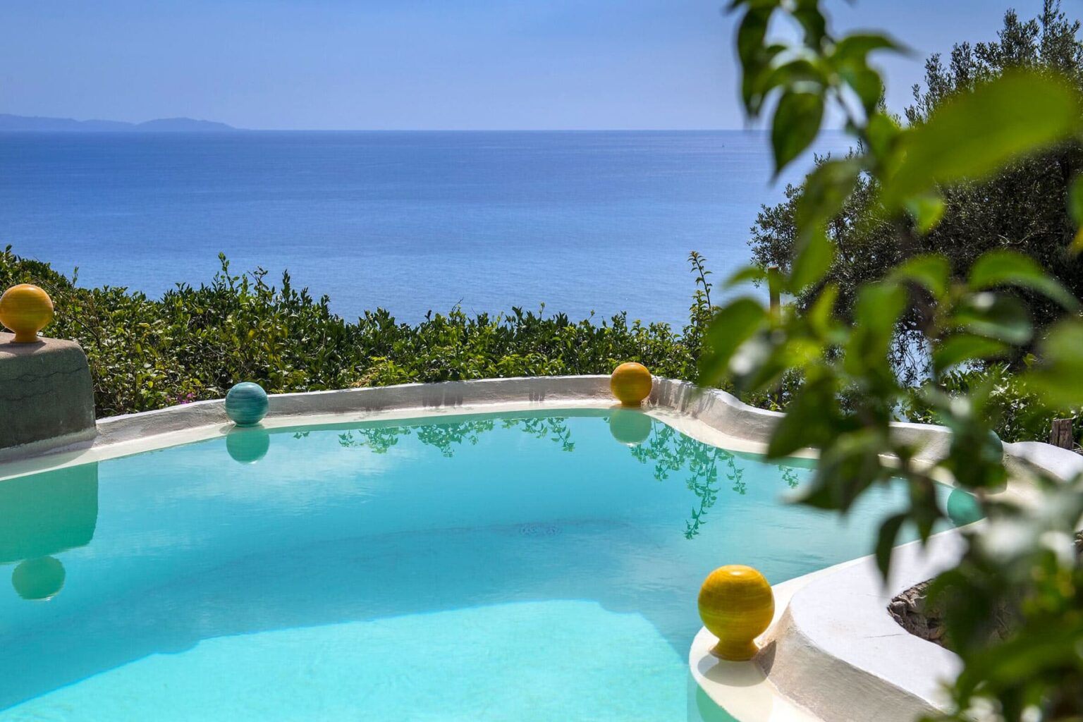 Scenic view of a clear blue swimming pool surrounded by lush greenery, with colorful floating orbs and a stunning ocean backdrop. Perfect for a relaxing getaway. | Only on SopranoVillas
