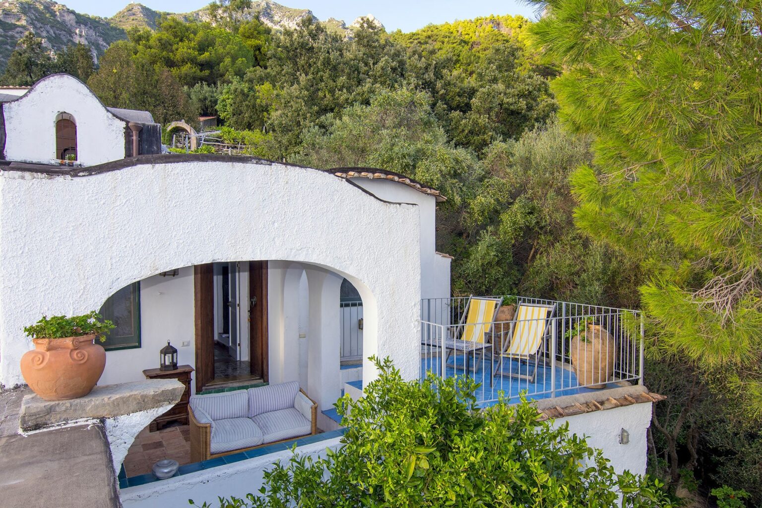 Charming white villa with an arched entrance, surrounded by lush greenery, featuring a private terrace with sun loungers and potted plants, ideal for relaxing outdoors. | Only on SopranoVillas