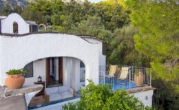 Charming white villa with an arched entrance, surrounded by lush greenery, featuring a private terrace with sun loungers and potted plants, ideal for relaxing outdoors.