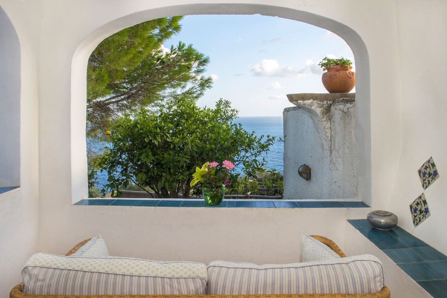Cozy interior view featuring a comfortable seating area with soft cushions, framed by an arched window overlooking a serene ocean landscape, lush greenery, and a potted plant, creating a tranquil and inviting atmosphere. | Only on SopranoVillas