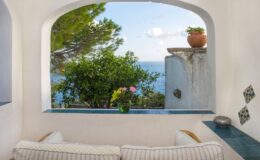 Cozy interior view featuring a comfortable seating area with soft cushions, framed by an arched window overlooking a serene ocean landscape, lush greenery, and a potted plant, creating a tranquil and inviting atmosphere.