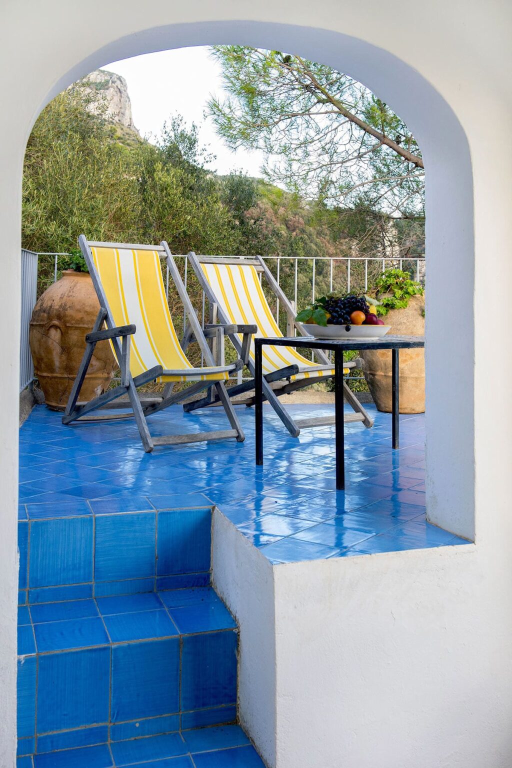 Cozy outdoor patio featuring two yellow-striped lounge chairs, a small black table with a bowl of fresh fruit, and terracotta pots with greenery, all set against a vibrant blue tiled floor. | Only on SopranoVillas