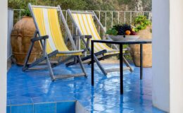 Cozy outdoor patio featuring two yellow-striped lounge chairs, a small black table with a bowl of fresh fruit, and terracotta pots with greenery, all set against a vibrant blue tiled floor.