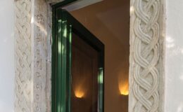 Intricately designed stone door frame featuring a braided pattern, leading to a warmly lit interior space with a green door.