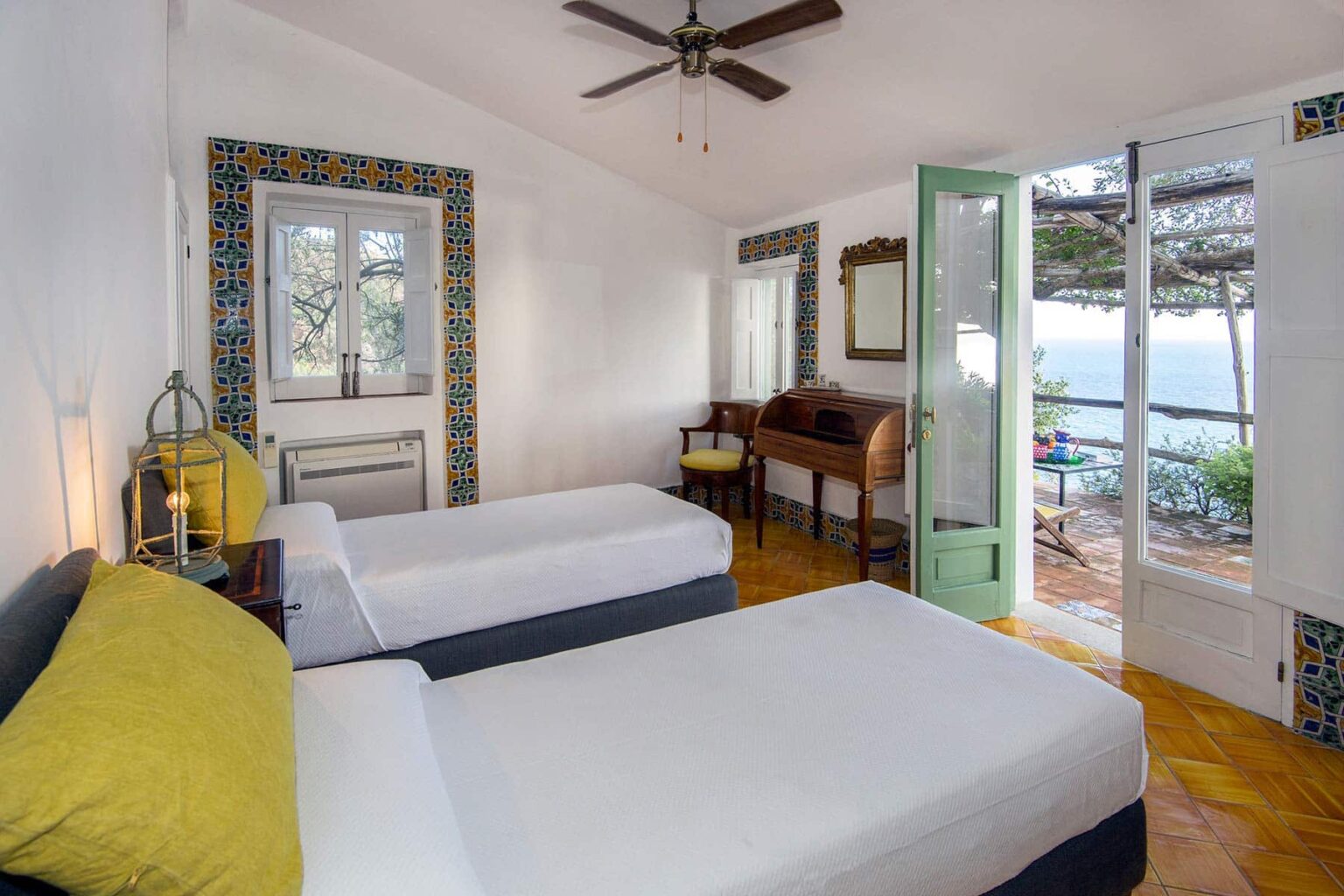 Bright and airy hotel room featuring two twin beds, a wooden desk, and large windows with ocean views. Decorated in light colors with a ceiling fan and stylish furnishings, this room offers a relaxing atmosphere for guests. | Only on SopranoVillas