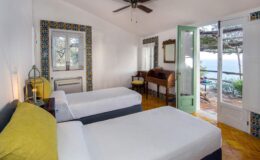 Bright and airy hotel room featuring two twin beds, a wooden desk, and large windows with ocean views. Decorated in light colors with a ceiling fan and stylish furnishings, this room offers a relaxing atmosphere for guests.