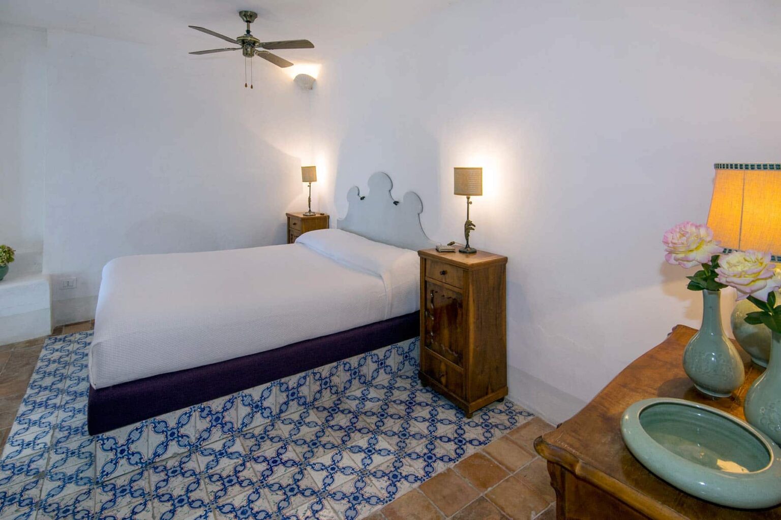 Cozy bedroom featuring a queen-sized bed with a minimalist design, wooden nightstands with lamps, a decorative blue patterned rug, and a stylish bedside table with a vase of flowers. The room is well-lit, highlighting the white walls and ceiling fan. | Only on SopranoVillas