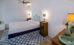 Cozy bedroom featuring a queen-sized bed with a minimalist design, wooden nightstands with lamps, a decorative blue patterned rug, and a stylish bedside table with a vase of flowers. The room is well-lit, highlighting the white walls and ceiling fan.