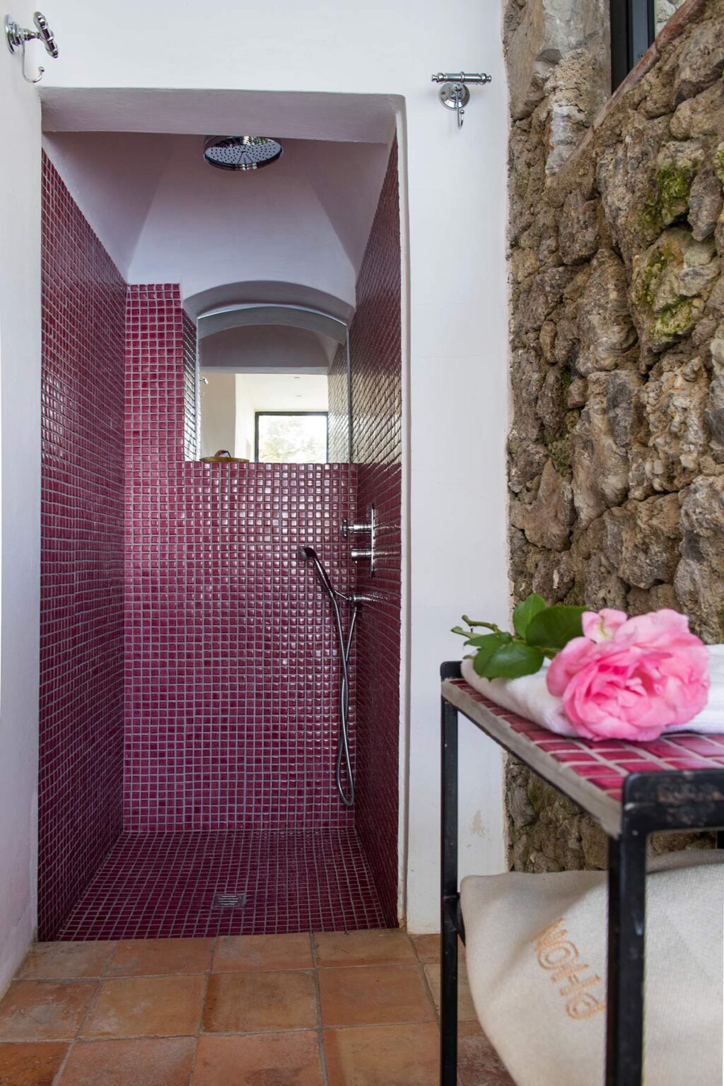 Modern shower with vibrant pink mosaic tiles and natural stone wall, featuring a sleek showerhead and decorative pink roses on a towel nearby, enhancing the spa-like ambiance. | Only on SopranoVillas