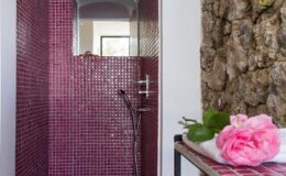 Modern shower with vibrant pink mosaic tiles and natural stone wall, featuring a sleek showerhead and decorative pink roses on a towel nearby, enhancing the spa-like ambiance.