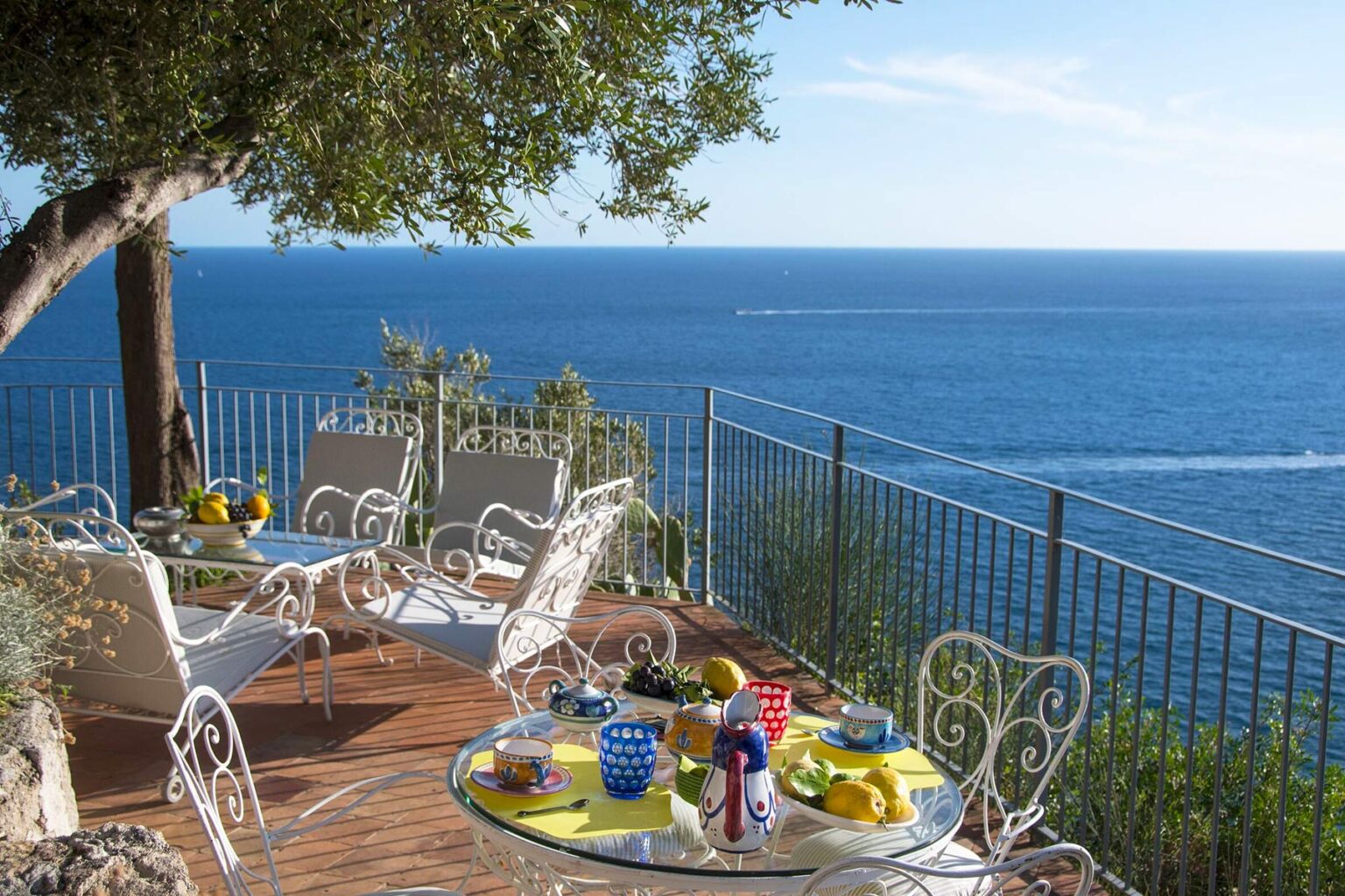 Scenic outdoor dining area overlooking the ocean, featuring white wrought iron furniture, colorful tableware, and a lush tree providing shade on a sunny day. | Only on SopranoVillas