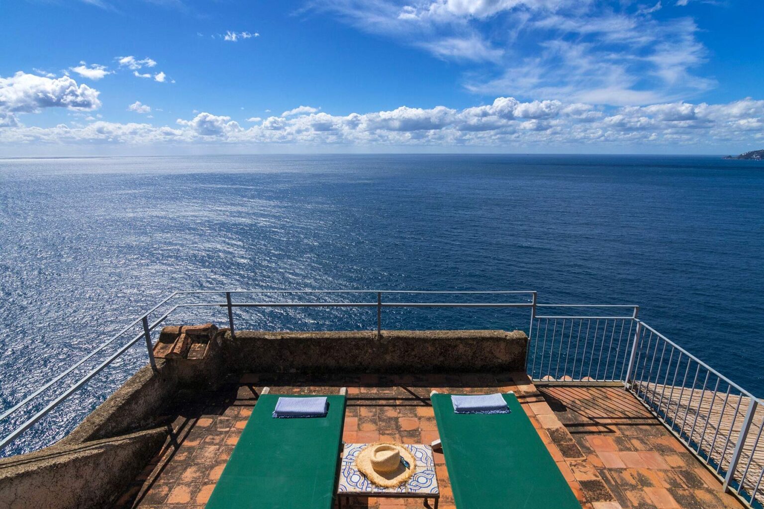 Panoramic view of a serene ocean from a terrace featuring two green sun loungers, perfect for relaxation under a bright blue sky with fluffy clouds. | Only on SopranoVillas