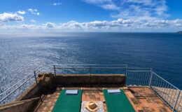 Panoramic view of a serene ocean from a terrace featuring two green sun loungers, perfect for relaxation under a bright blue sky with fluffy clouds.