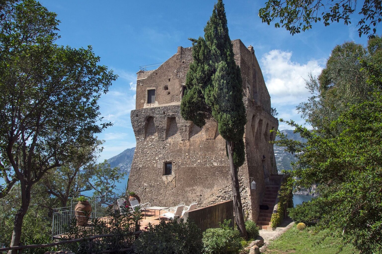 Historic stone tower surrounded by lush greenery and trees, overlooking a scenic landscape with mountains and a body of water. Ideal for travel and architectural history enthusiasts. | Only on SopranoVillas