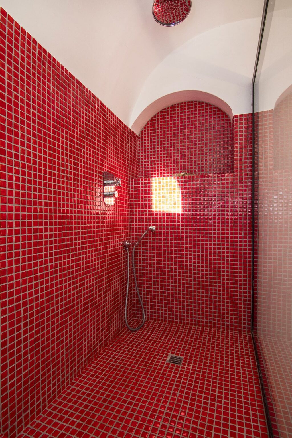 Interior view of a modern shower featuring vibrant red mosaic tiles, a handheld showerhead, and a window allowing natural light to enter the space. | Only on SopranoVillas