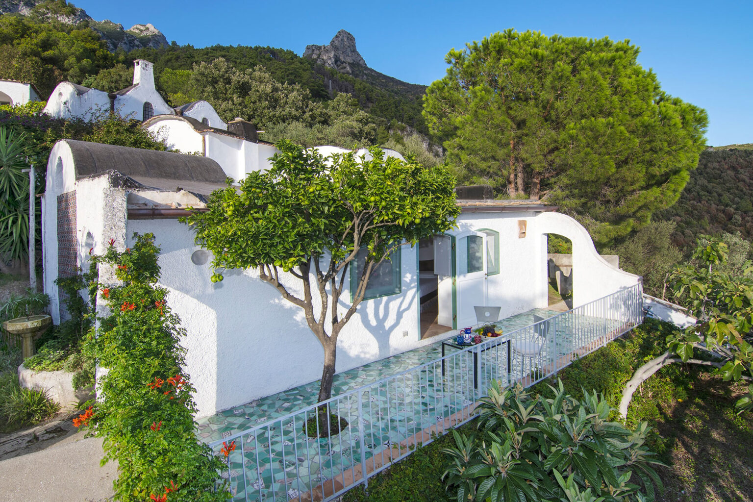 Charming white cottage surrounded by lush greenery and a scenic landscape, featuring a tree in the foreground and mountains in the background. Ideal for a tranquil getaway. | Only on SopranoVillas