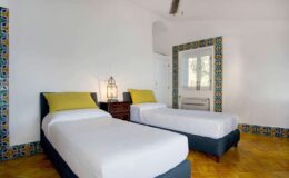Bright and airy bedroom featuring two twin beds with yellow accent pillows, wooden flooring, and decorative tiled walls, ideal for a comfortable stay.
