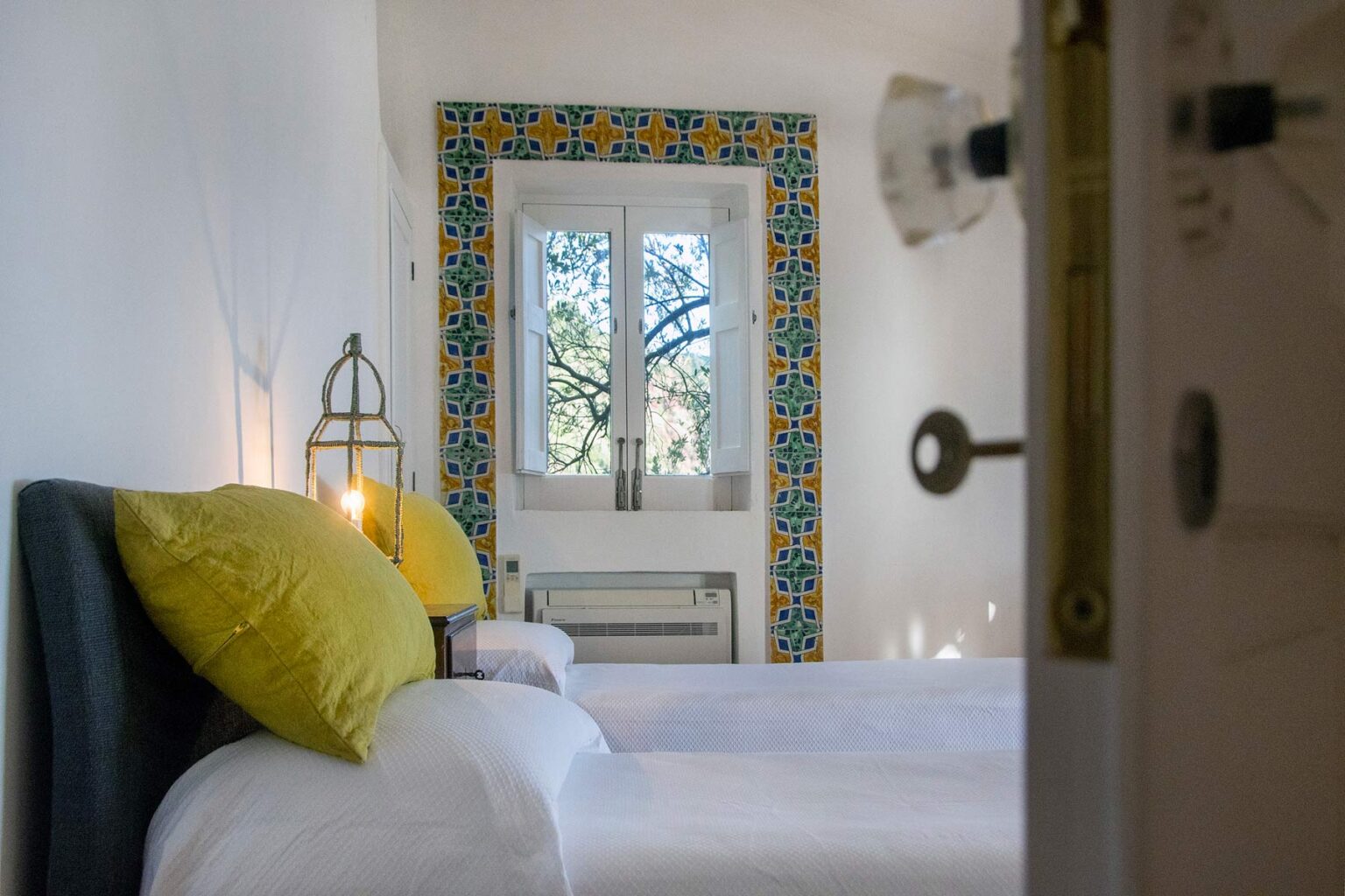 Bright and inviting bedroom featuring two twin beds with yellow accent pillows, a decorative tiled window frame, and natural light streaming in, creating a cozy atmosphere. | Only on SopranoVillas
