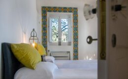 Bright and inviting bedroom featuring two twin beds with yellow accent pillows, a decorative tiled window frame, and natural light streaming in, creating a cozy atmosphere.