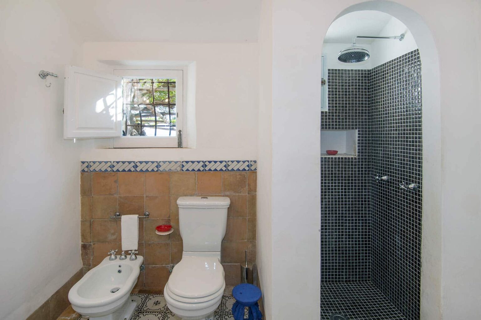 Modern bathroom featuring a white toilet and bidet, with a separate shower area finished in black tiles. The space includes a window for natural light and decorative tile accents. | Only on SopranoVillas