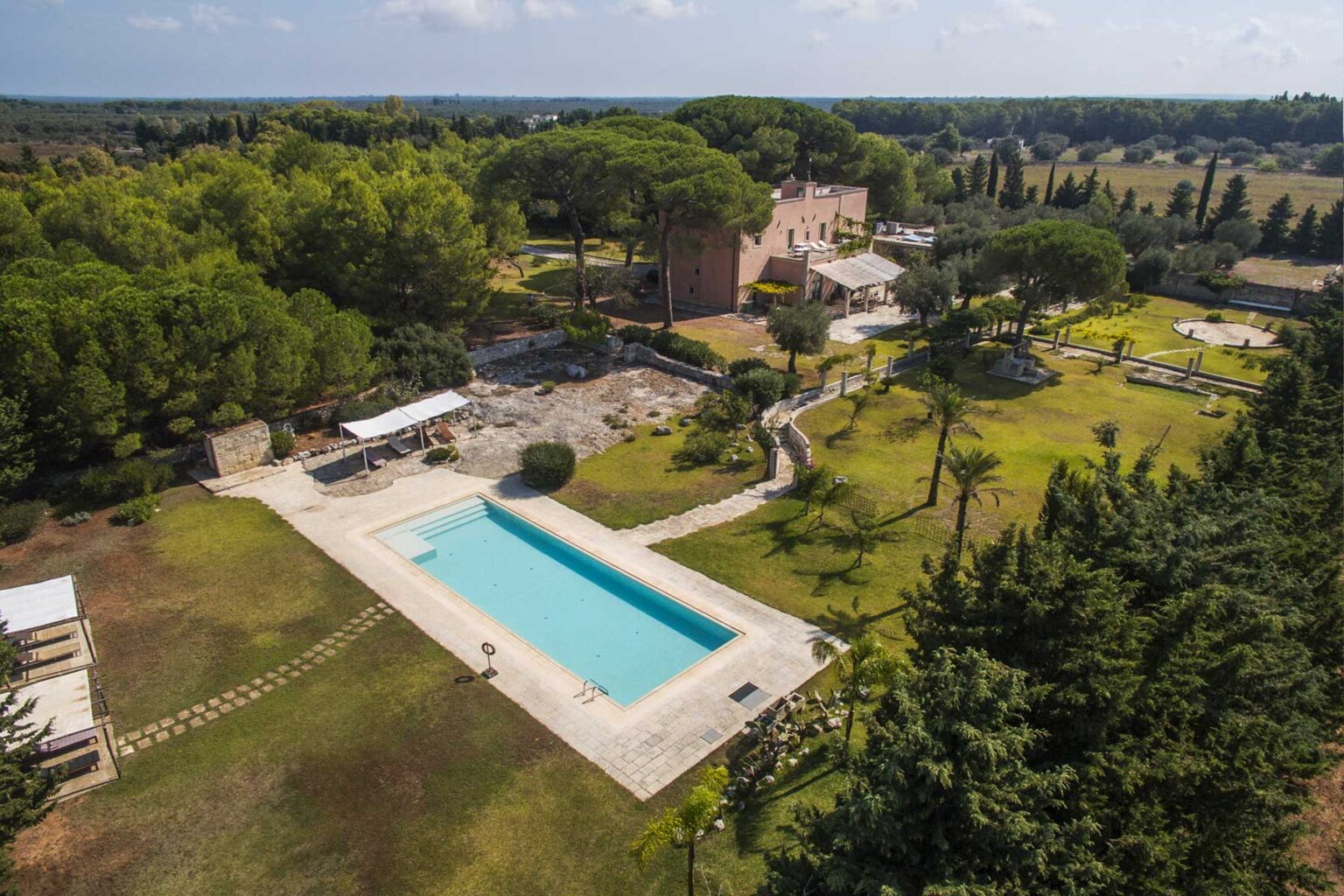 Aerial view of a luxurious villa with a large swimming pool surrounded by lush greenery and landscaped gardens, featuring outdoor seating areas and a scenic countryside backdrop. | Only on SopranoVillas
