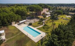 Aerial view of a luxurious villa with a large swimming pool surrounded by lush greenery and landscaped gardens, featuring outdoor seating areas and a scenic countryside backdrop.