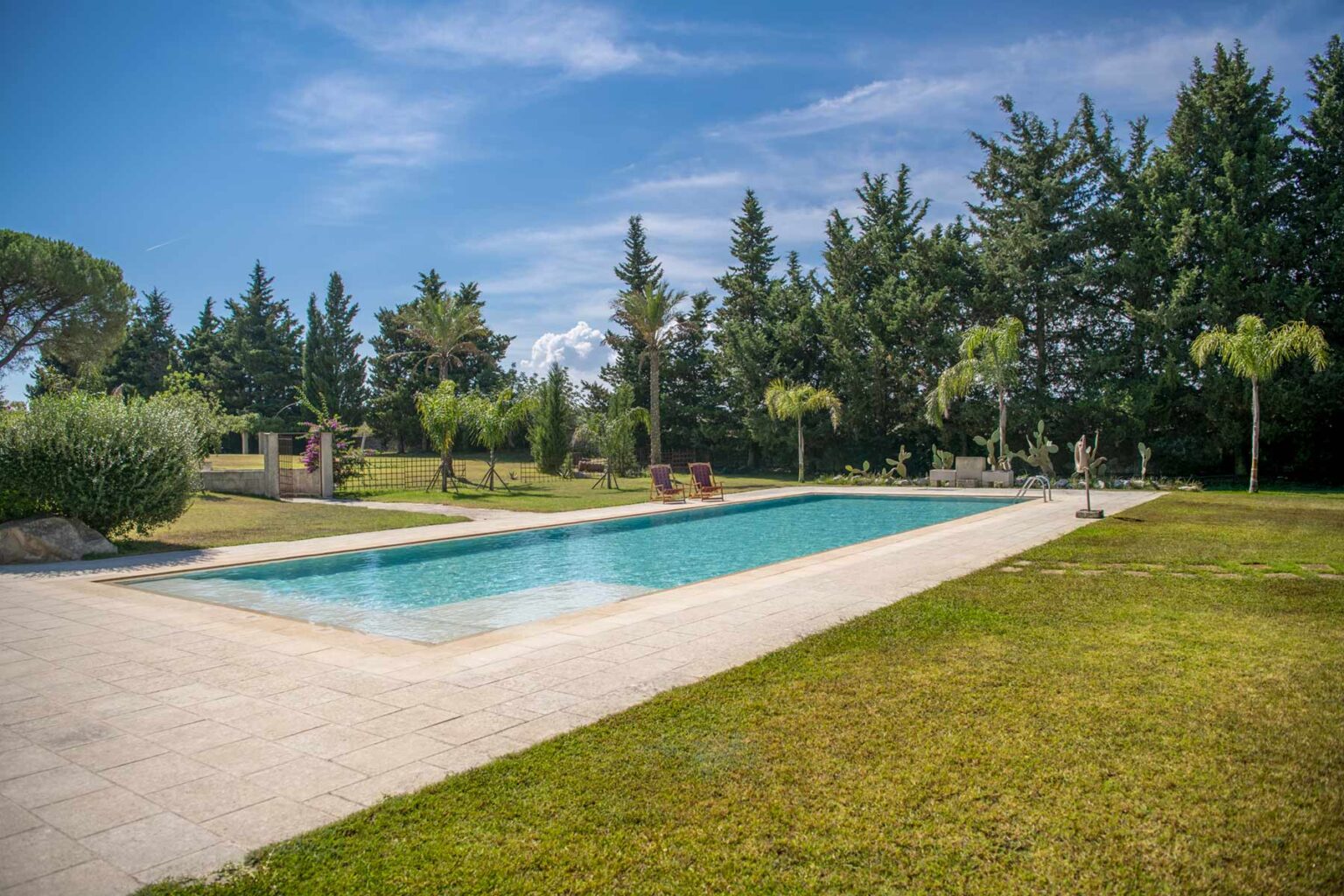 Scenic outdoor swimming pool surrounded by lush greenery and tall trees under a clear blue sky, ideal for relaxation and leisure activities. | Only on SopranoVillas