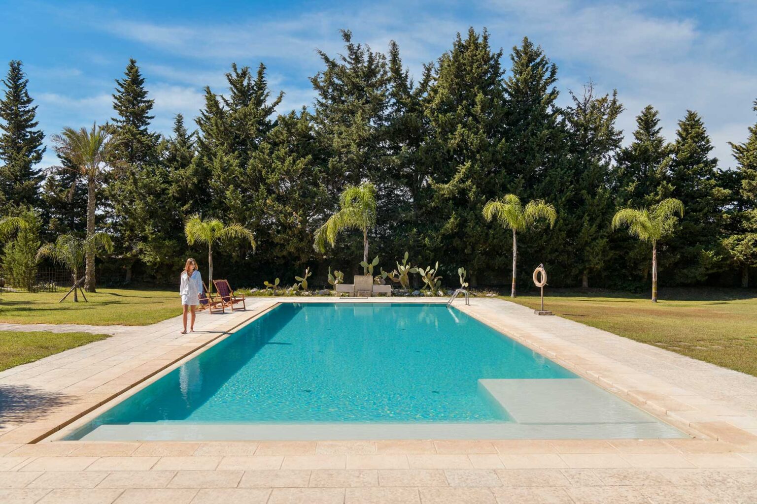 A serene outdoor swimming pool surrounded by lush greenery and palm trees, with a person relaxing by the poolside. Ideal for summer retreats and relaxation. | Only on SopranoVillas