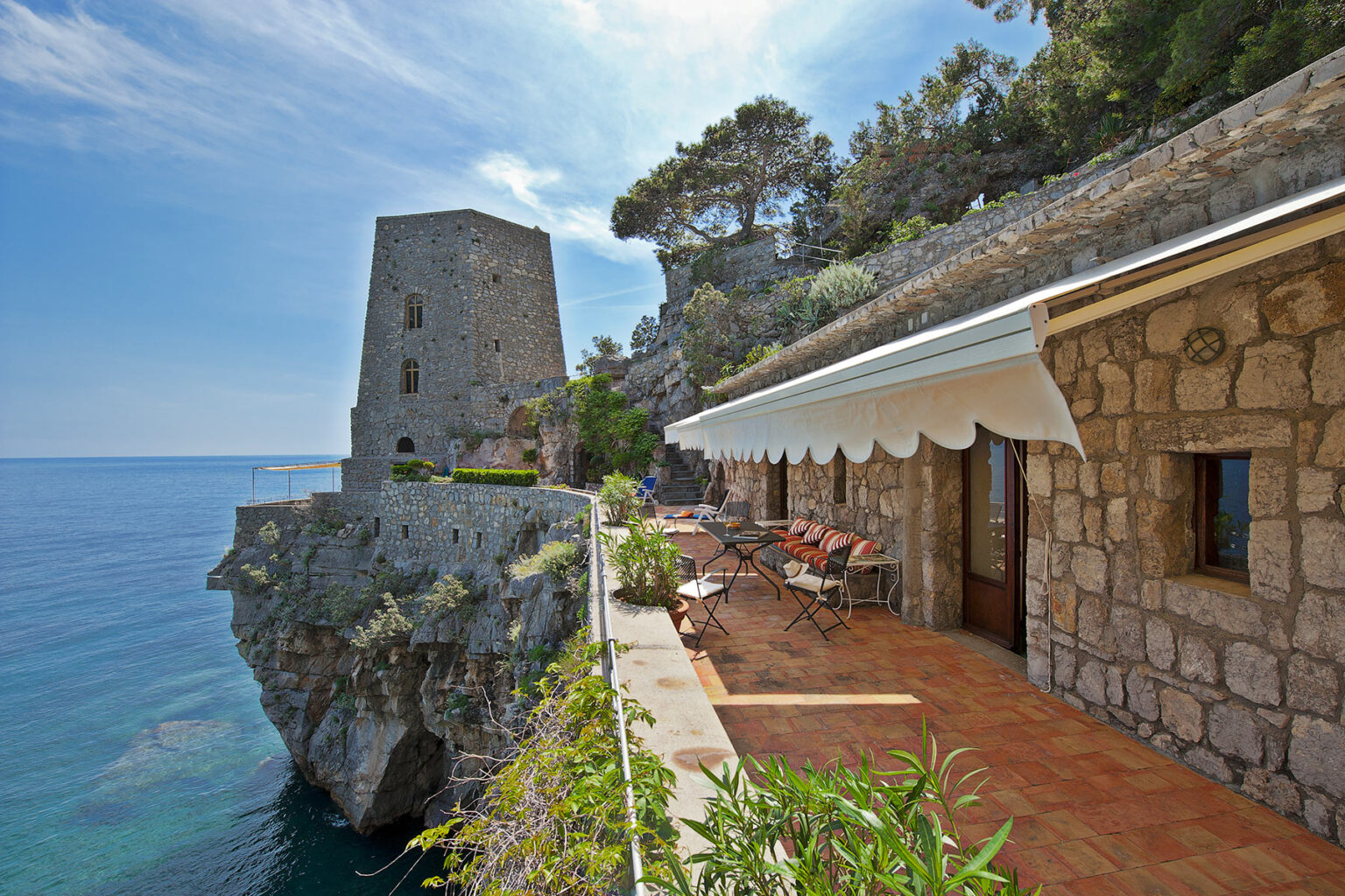 Scenic coastal view featuring a historic stone tower and a charming villa with outdoor seating, surrounded by lush greenery and overlooking the sea under a clear blue sky. | Only on SopranoVillas
