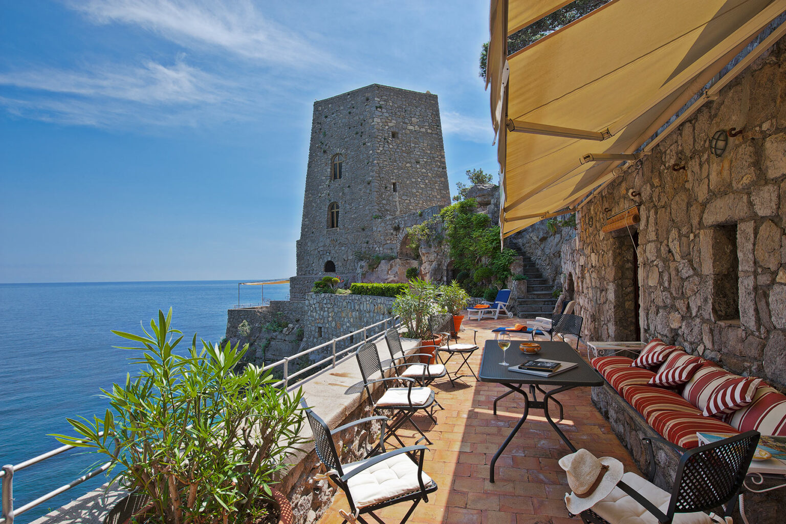 Coastal view of a stone terrace with seating, overlooking the sea and an ancient tower, under a clear blue sky. Ideal for vacation rentals or seaside retreats. | Only on SopranoVillas