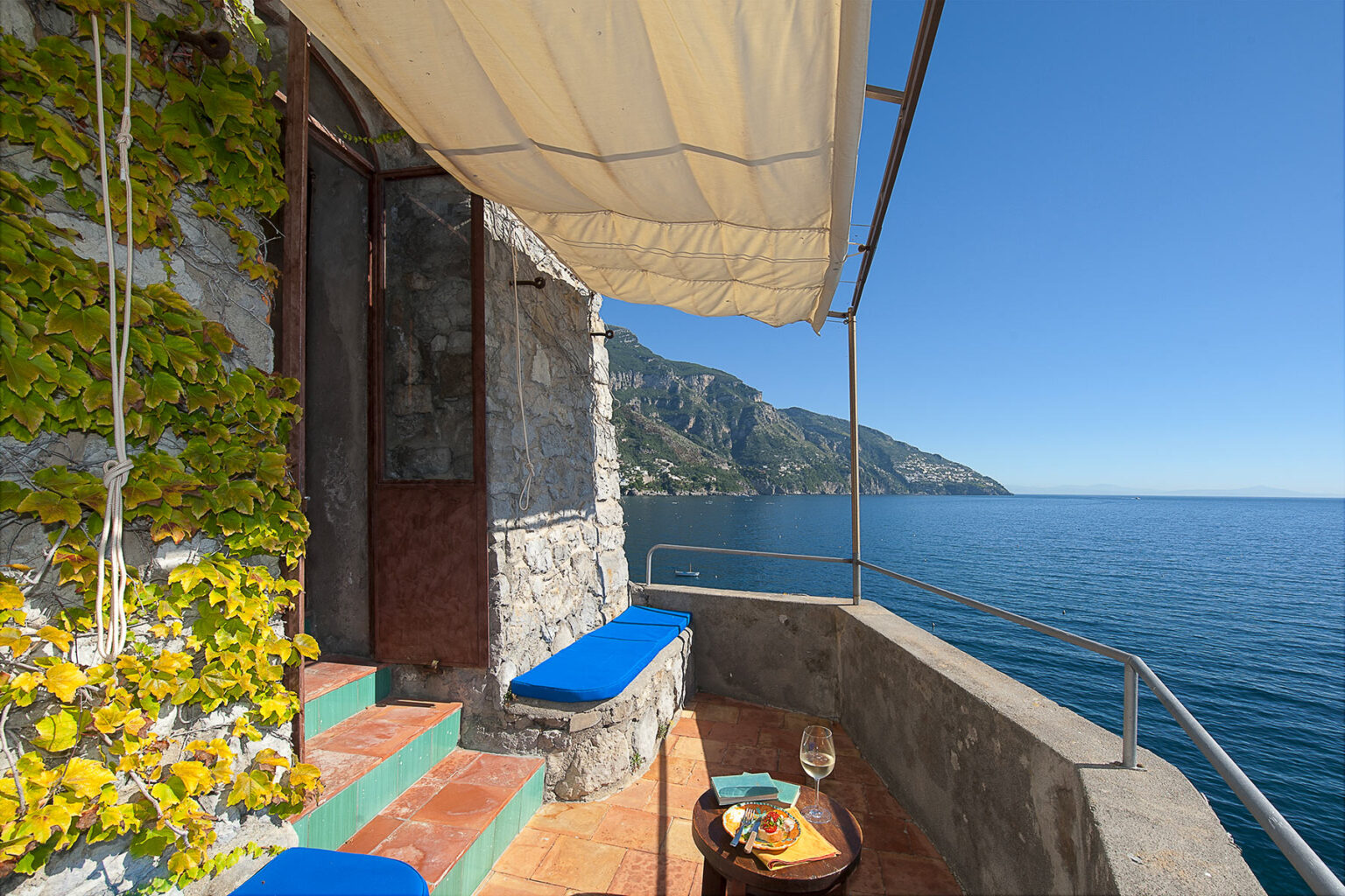 Scenic coastal terrace featuring blue seating, a stone wall, and a stunning view of the ocean and mountains under a clear blue sky. Perfect for relaxing and enjoying the seaside atmosphere. | Only on SopranoVillas