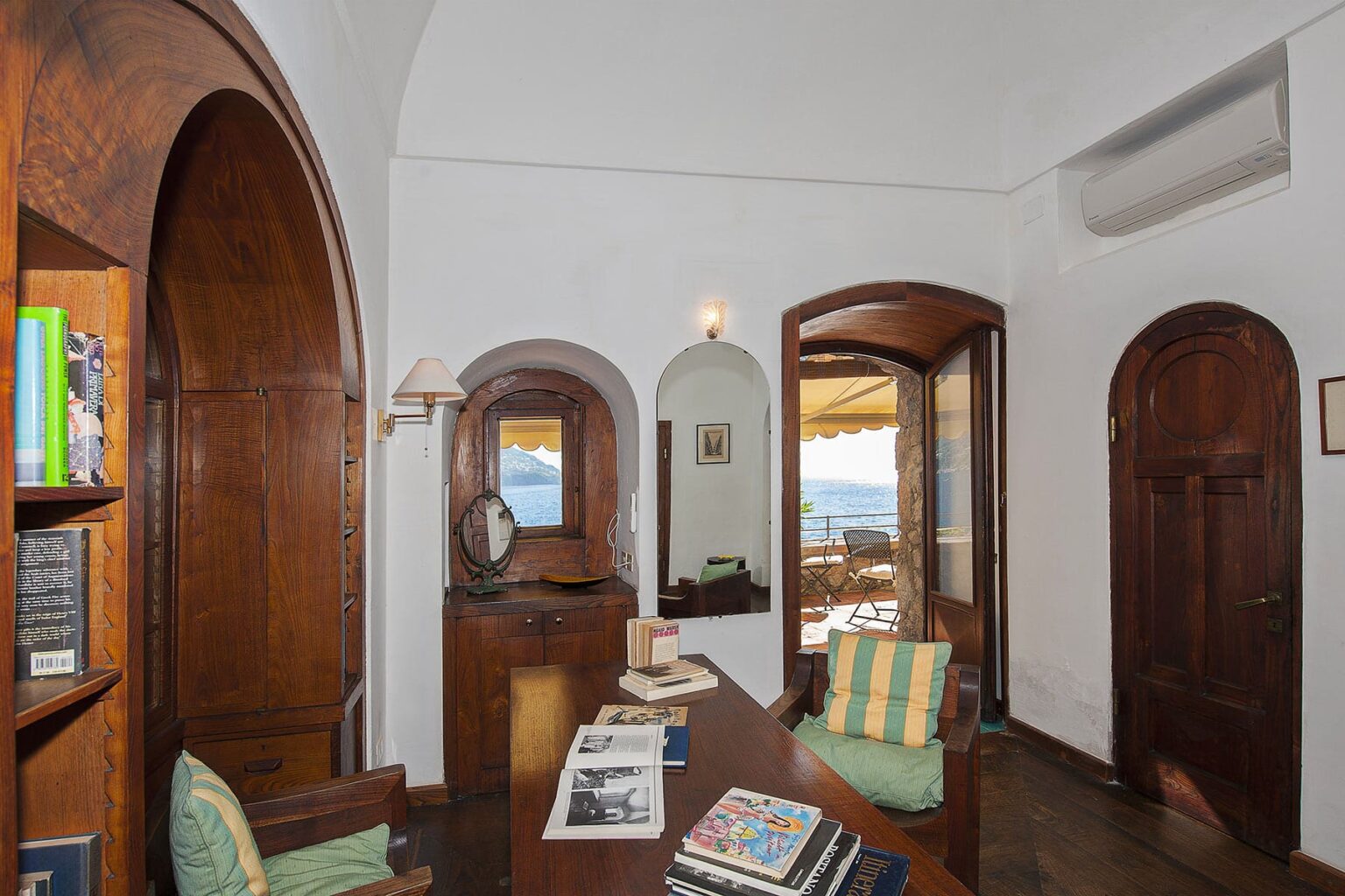 Cozy interior of a stylish office featuring wooden arches, a desk with magazines, a comfortable chair, and a view through arched windows. The room has a bright, airy feel with natural light streaming in. | Only on SopranoVillas