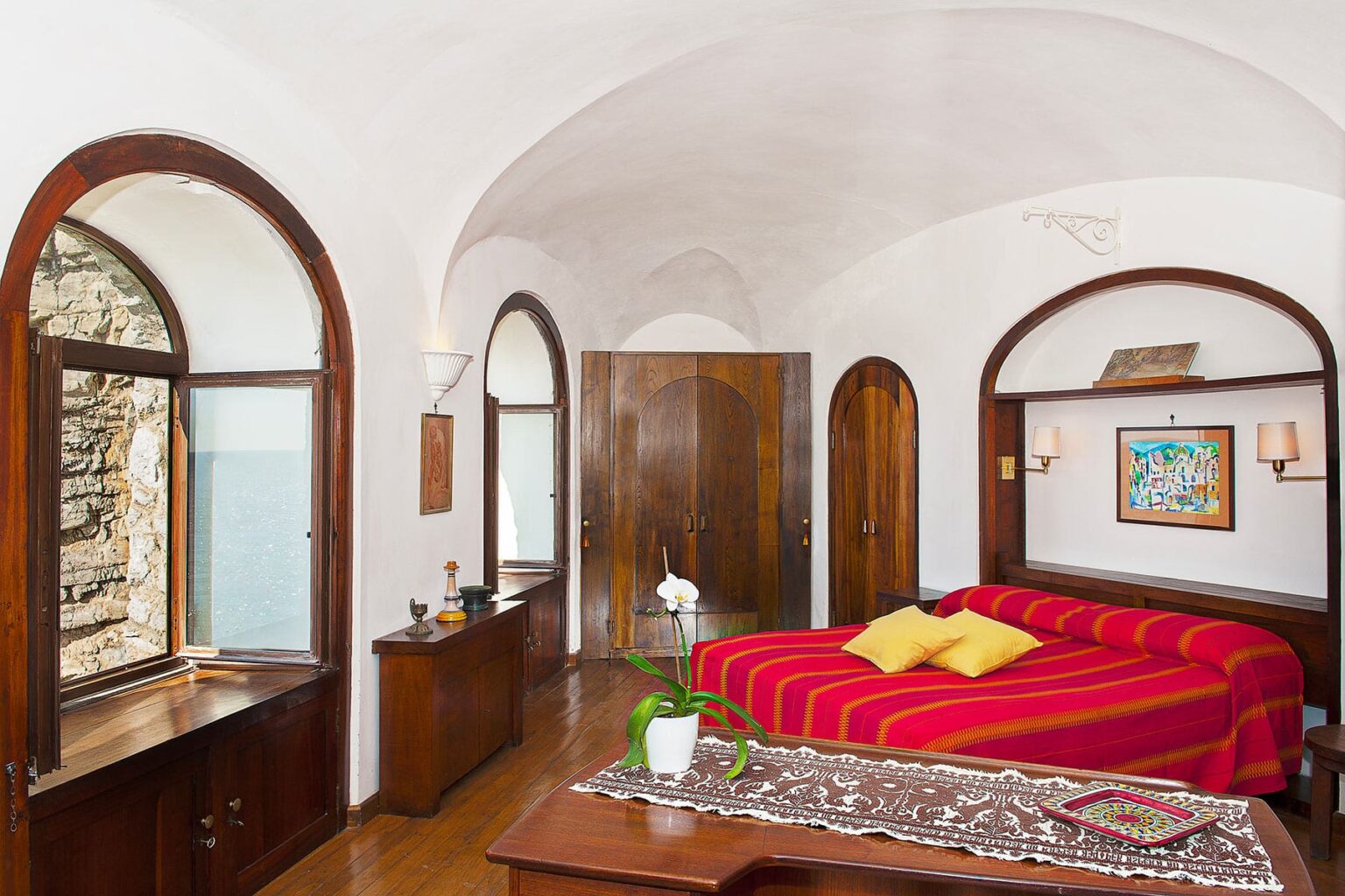 Cozy bedroom with a red-striped bedspread, wooden furniture, and large arched windows showcasing a scenic view. The room features a warm color palette and decorative elements, creating a welcoming atmosphere. | Only on SopranoVillas