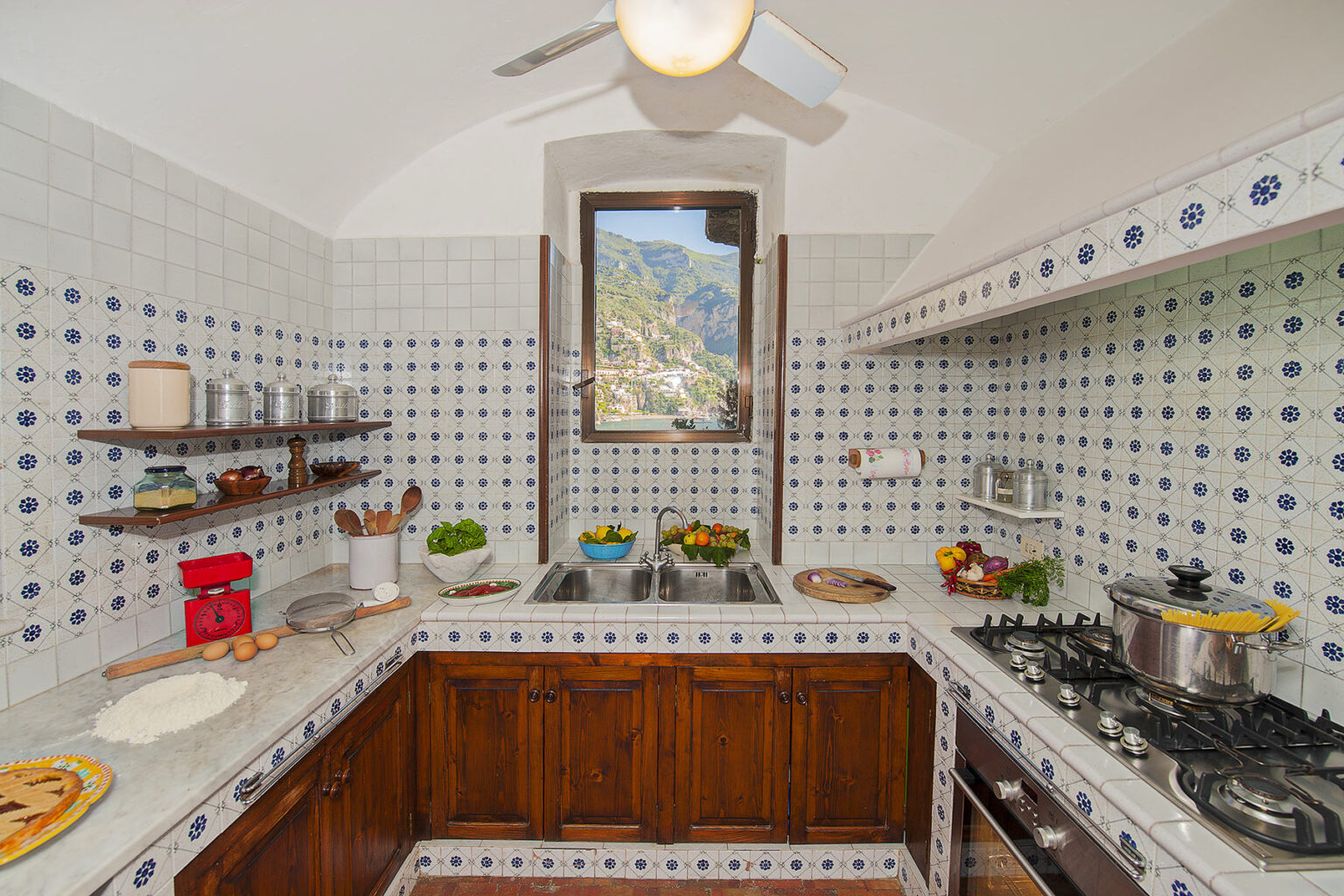 Bright and inviting kitchen featuring traditional blue and white tiled walls, wooden cabinetry, and modern appliances. Natural light floods in through a window with a scenic view of mountains, while fresh ingredients and cooking utensils are neatly arranged on the countertops. | Only on SopranoVillas