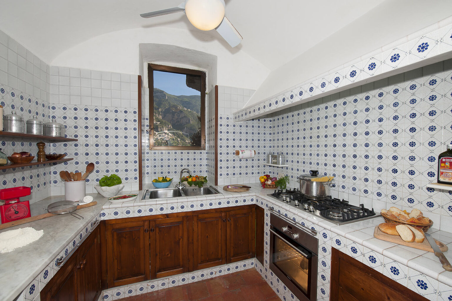 Bright and airy kitchen featuring white tiled walls with blue patterns, wooden cabinetry, a gas stove, and a window with a mountain view. Fresh ingredients are displayed on the countertop, enhancing the inviting atmosphere. | Only on SopranoVillas