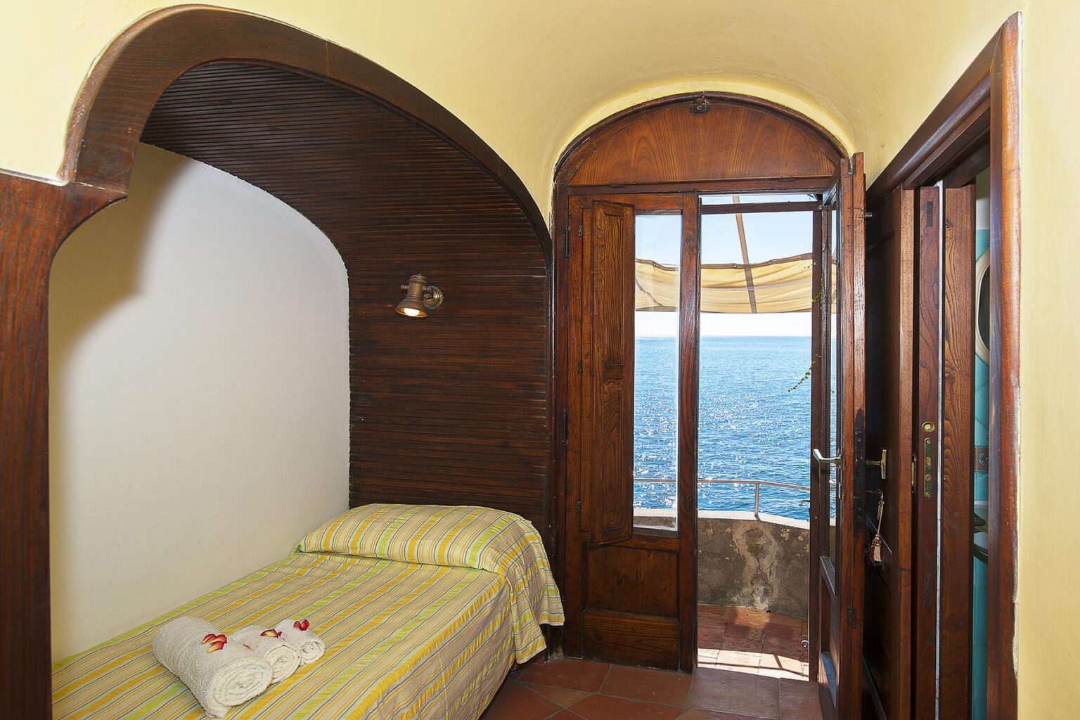 Cozy bedroom with a single bed and wooden accents, featuring a large window that opens to a stunning ocean view. The room is decorated with yellow bedding and has an inviting atmosphere, perfect for relaxation. | Only on SopranoVillas